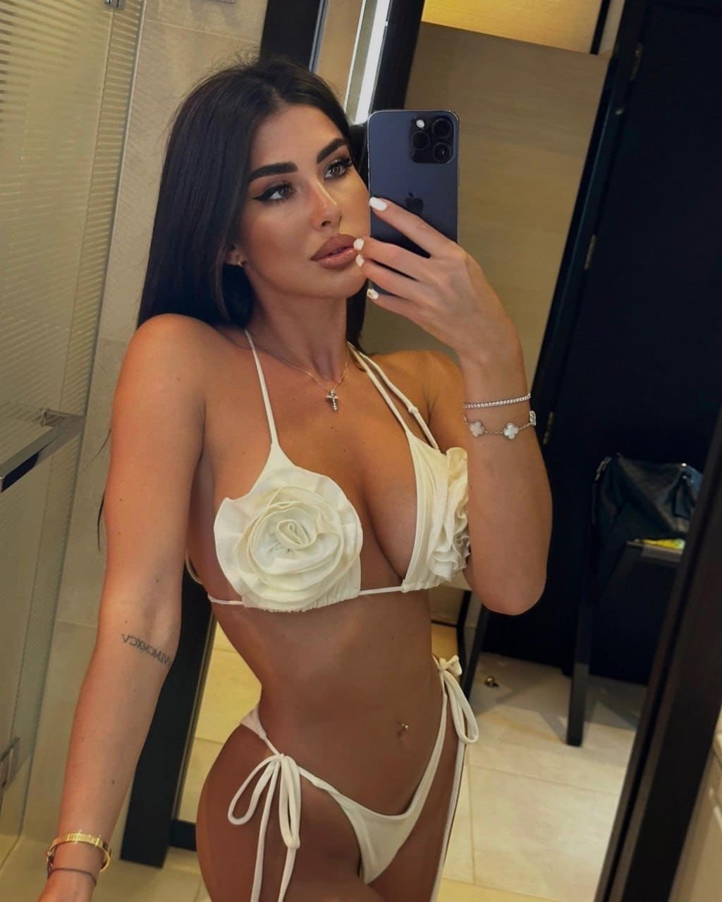 Mirror selfie of a woman in a white floral bikini holding a smartphone, standing in a tiled room with a glass door and black bag in the background.