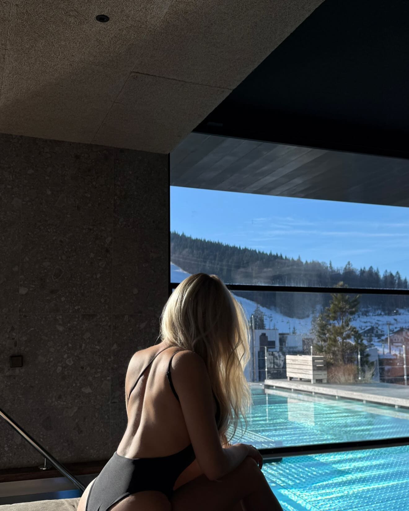 Back view of a blonde woman in a black swimsuit sitting by an indoor pool, looking out at a snowy landscape through large windows.