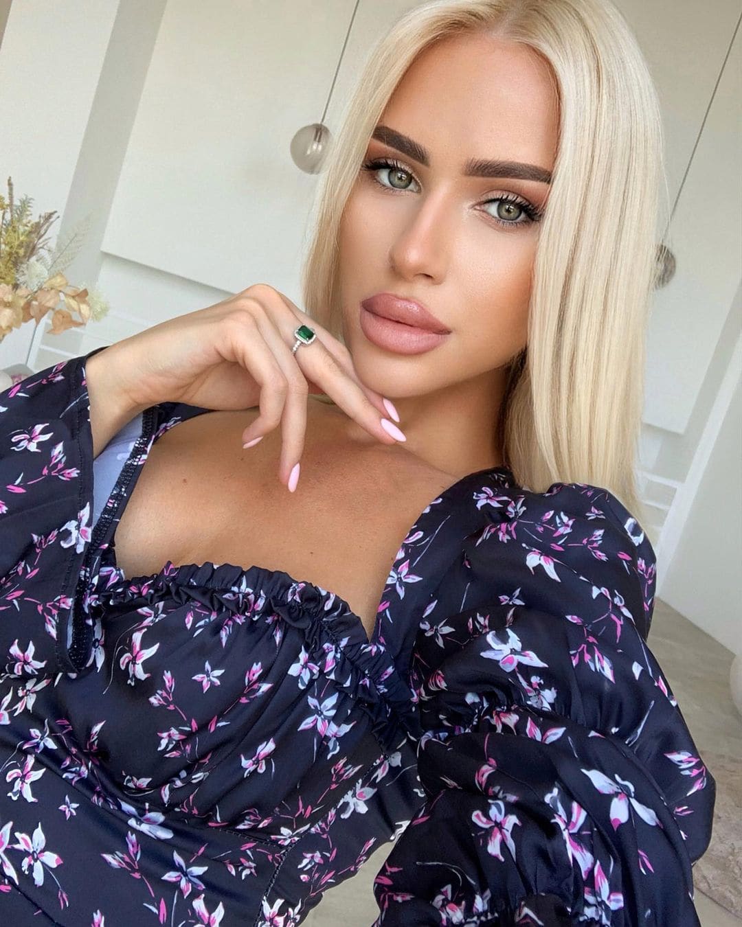 Indoors, she wears a dark floral top with puffed sleeves, raising a hand with a green-stone ring and pink nails near her chin.
