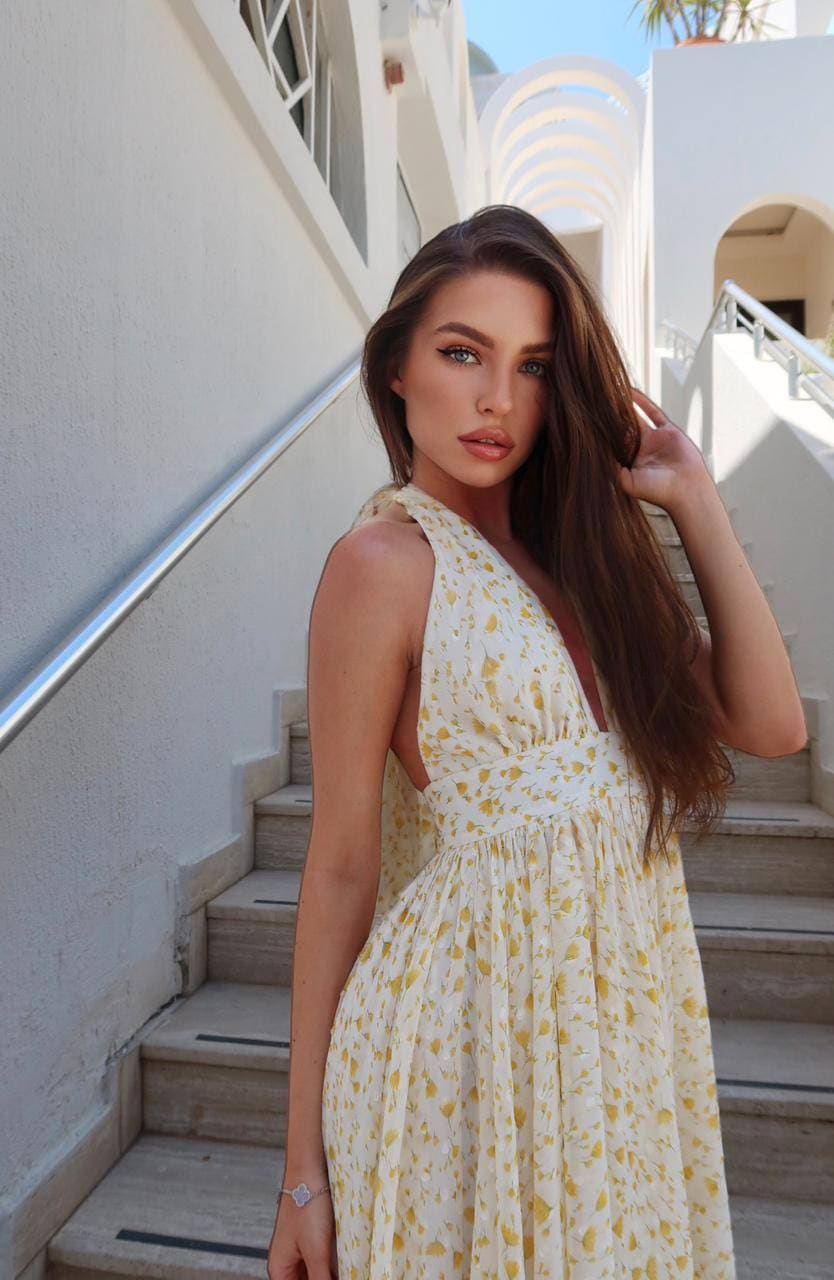Standing on stone steps, a woman in a white floral V-neck dress touches her hair while looking toward the camera in daylight.