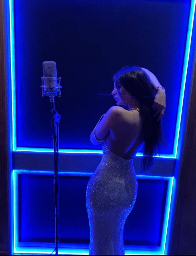 A woman in a sparkling silver backless gown stands in profile at a studio microphone, hand to head, lit by blue neon panels.