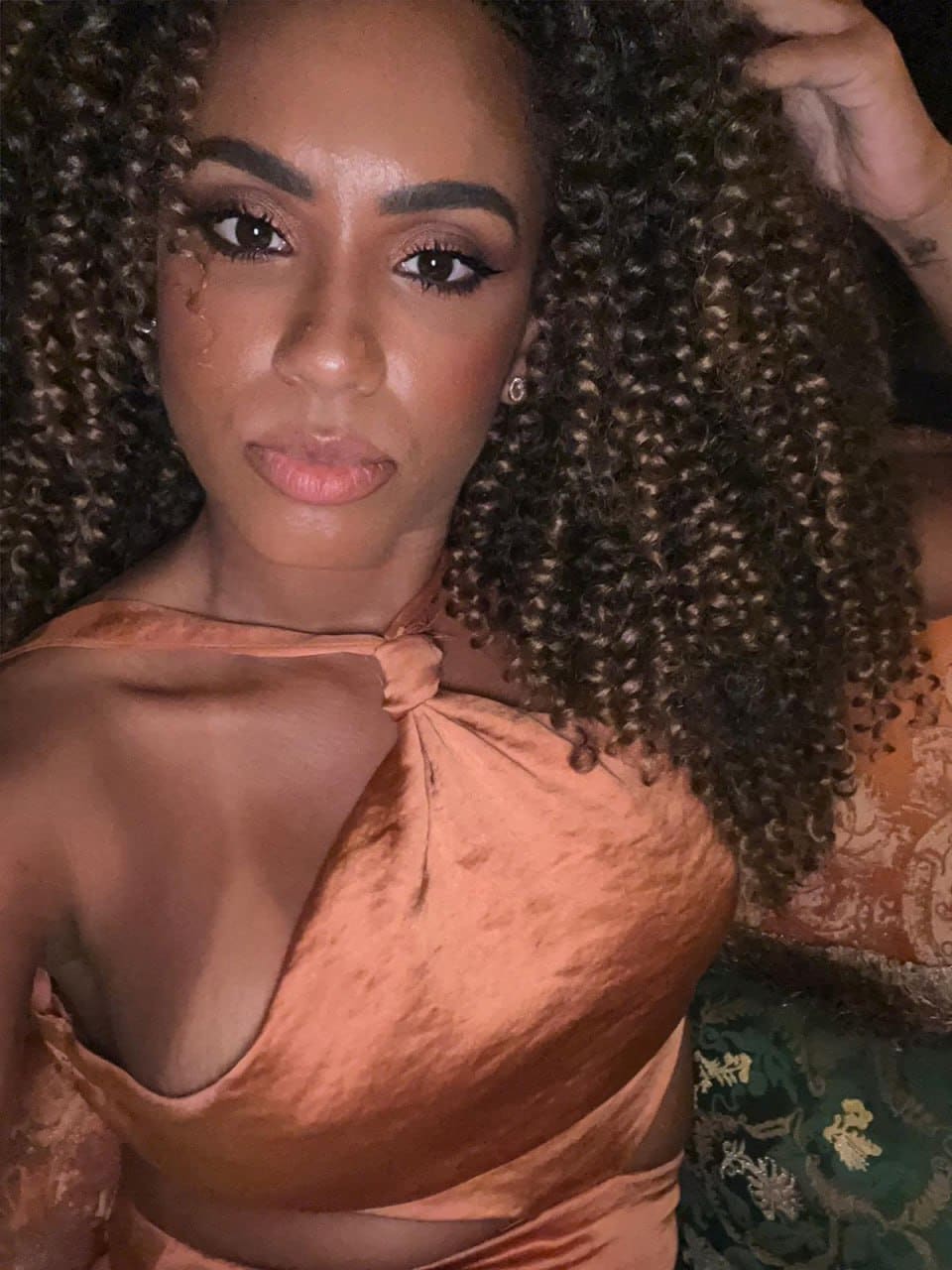 A close-up portrait of a woman in a peach satin halter top with bare shoulders, posing indoors against a patterned green and gold fabric background.