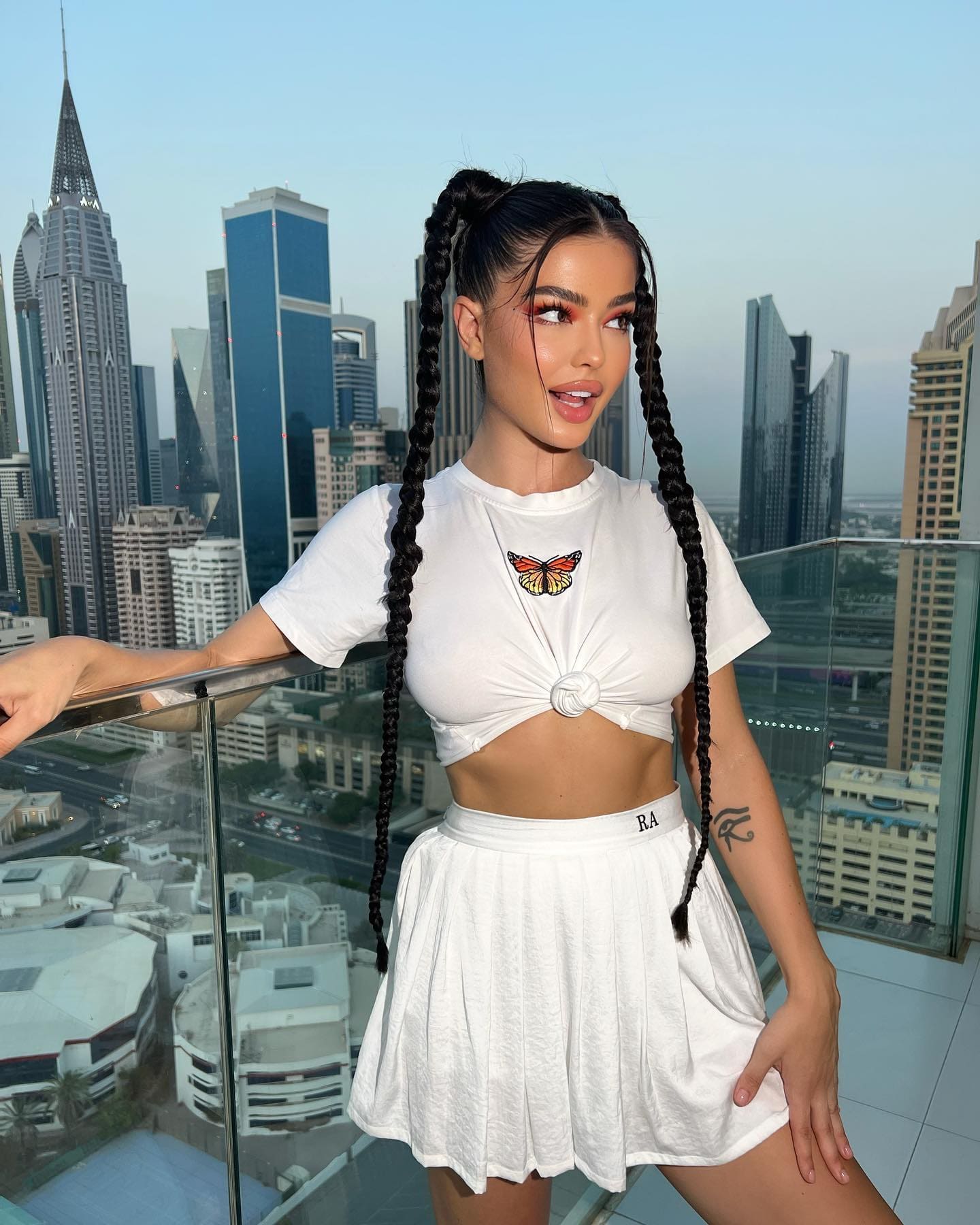 A woman with braided hair wears a white butterfly crop top and pleated skirt on a balcony, hand on glass railing.