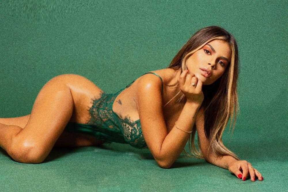 Lying on her side on a green surface, she wears a green lace bodysuit with her hand near her chin.
