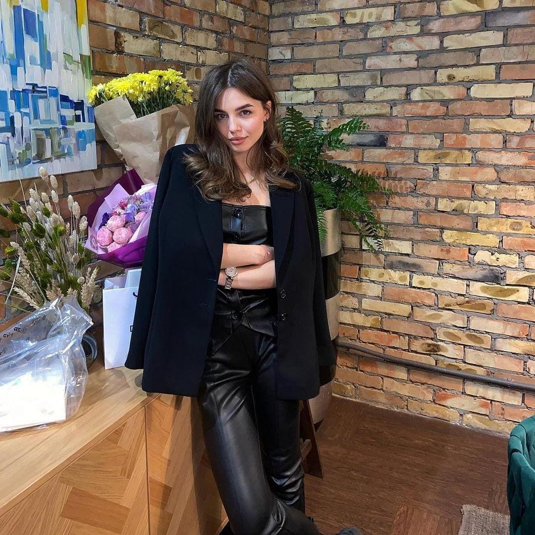 Woman in a black blazer and leather outfit stands with arms crossed near a brick wall and wooden counter with plants.
