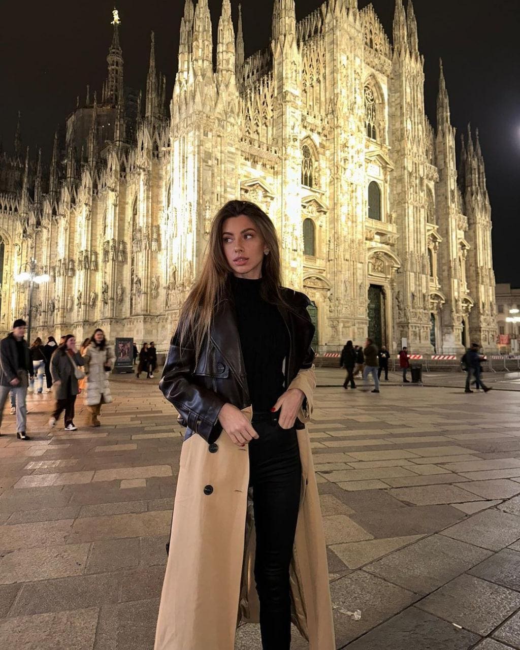Leila stands in a paved plaza at night before an illuminated Gothic cathedral, wearing a black turtleneck, pants, leather jacket, and beige coat.