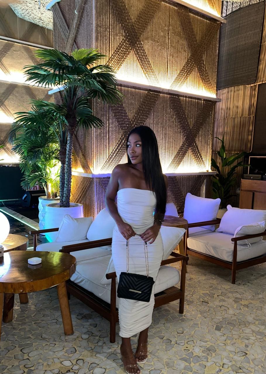 Woman in a white strapless midi dress holds a black handbag in a lounge with white chairs and plants.