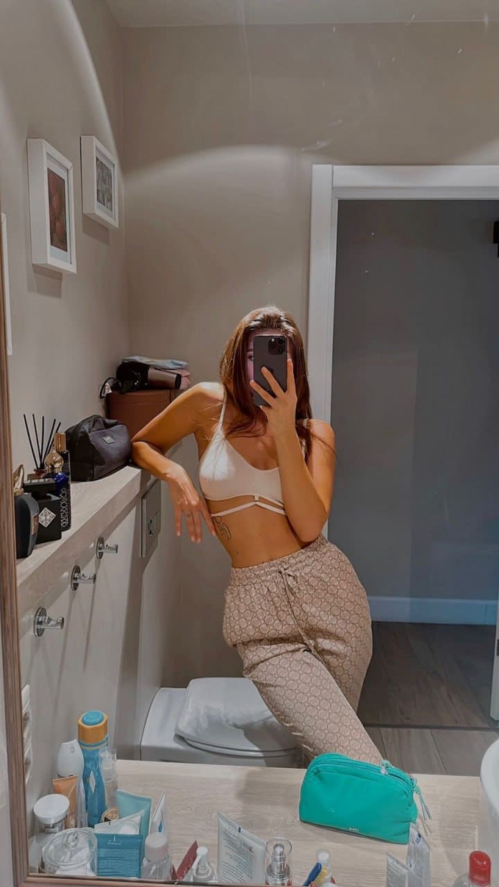 A woman stands in a bathroom wearing a white strappy bralette and beige sweatpants, holding a phone over her face.