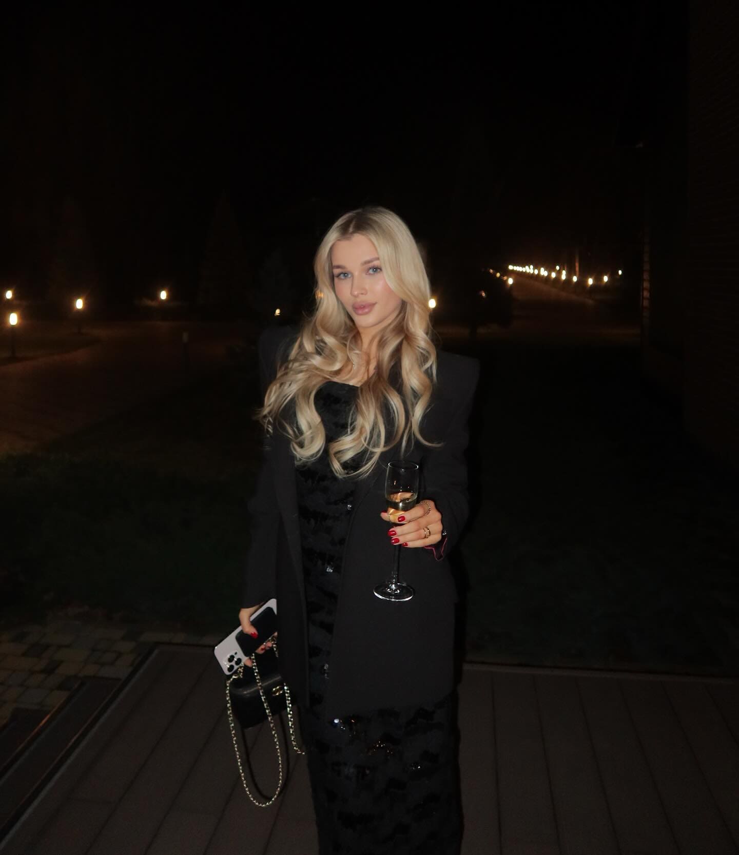 Madeleine stands on a wooden deck at night in a black blazer and patterned dress, holding a glass of white wine and a phone.