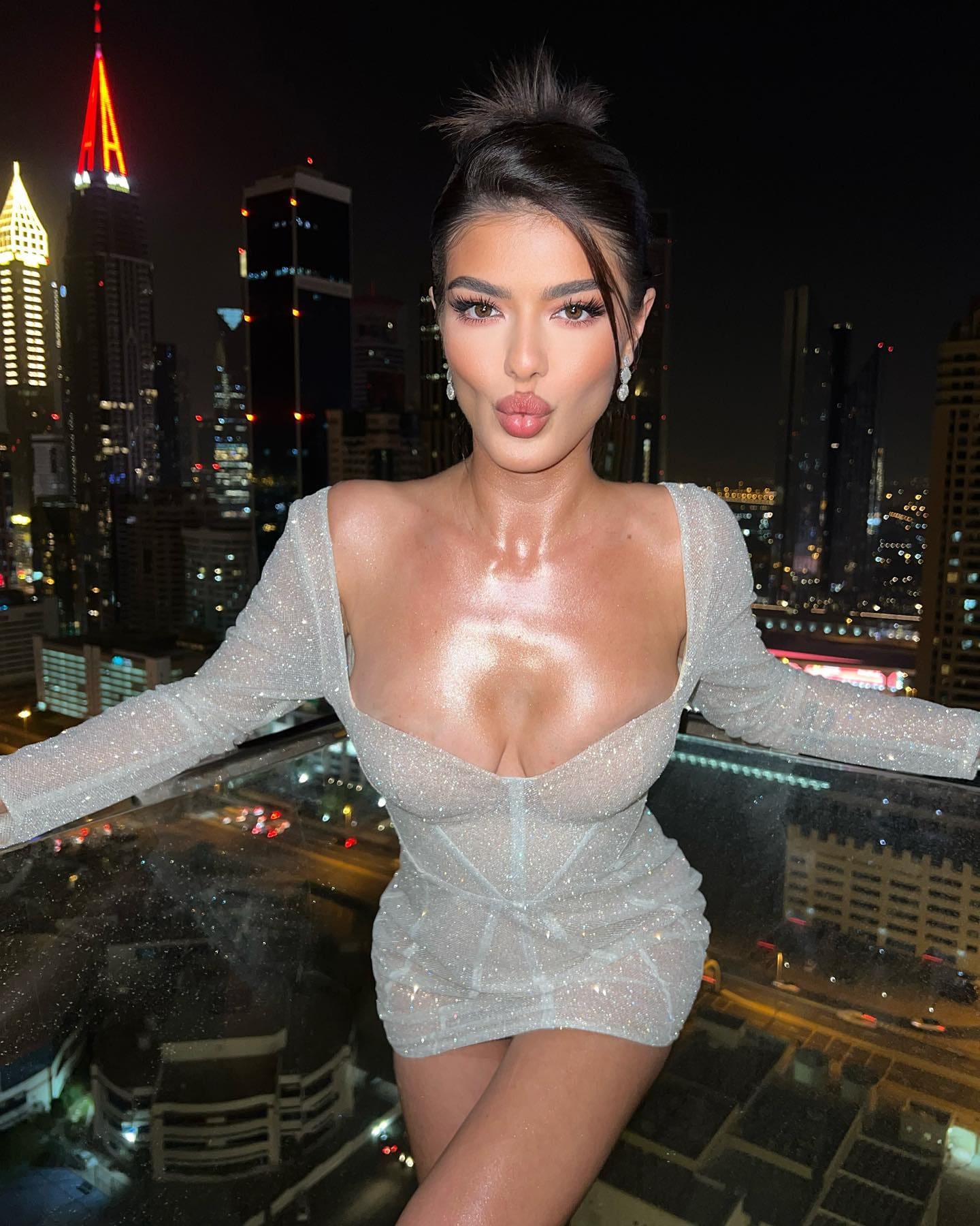 A woman in a sparkly silver mini dress poses with arms outstretched on a rooftop, illuminated skyscrapers at night.
