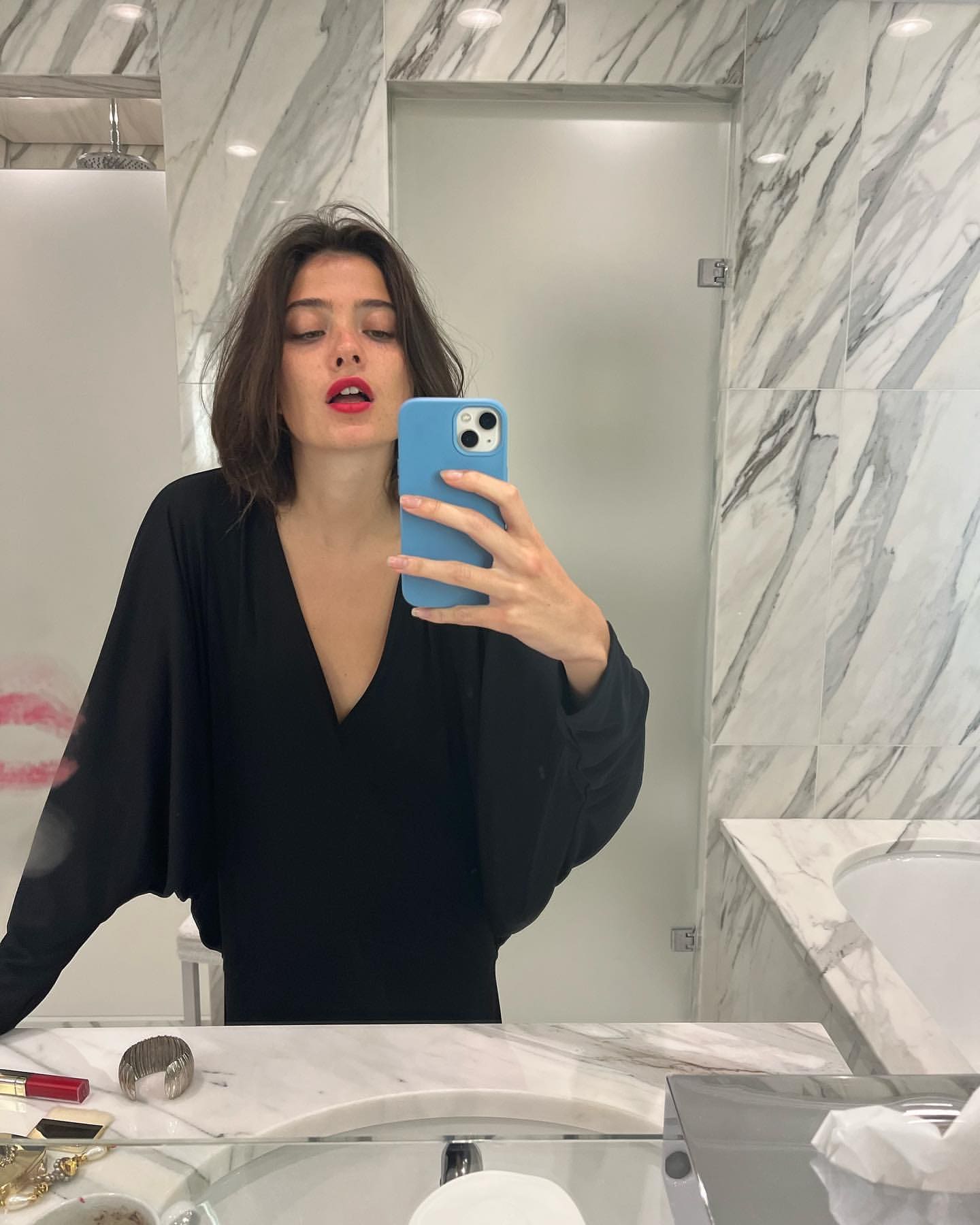 A mirror selfie in a marble bathroom shows her in a black V-neck top, holding a blue phone case.