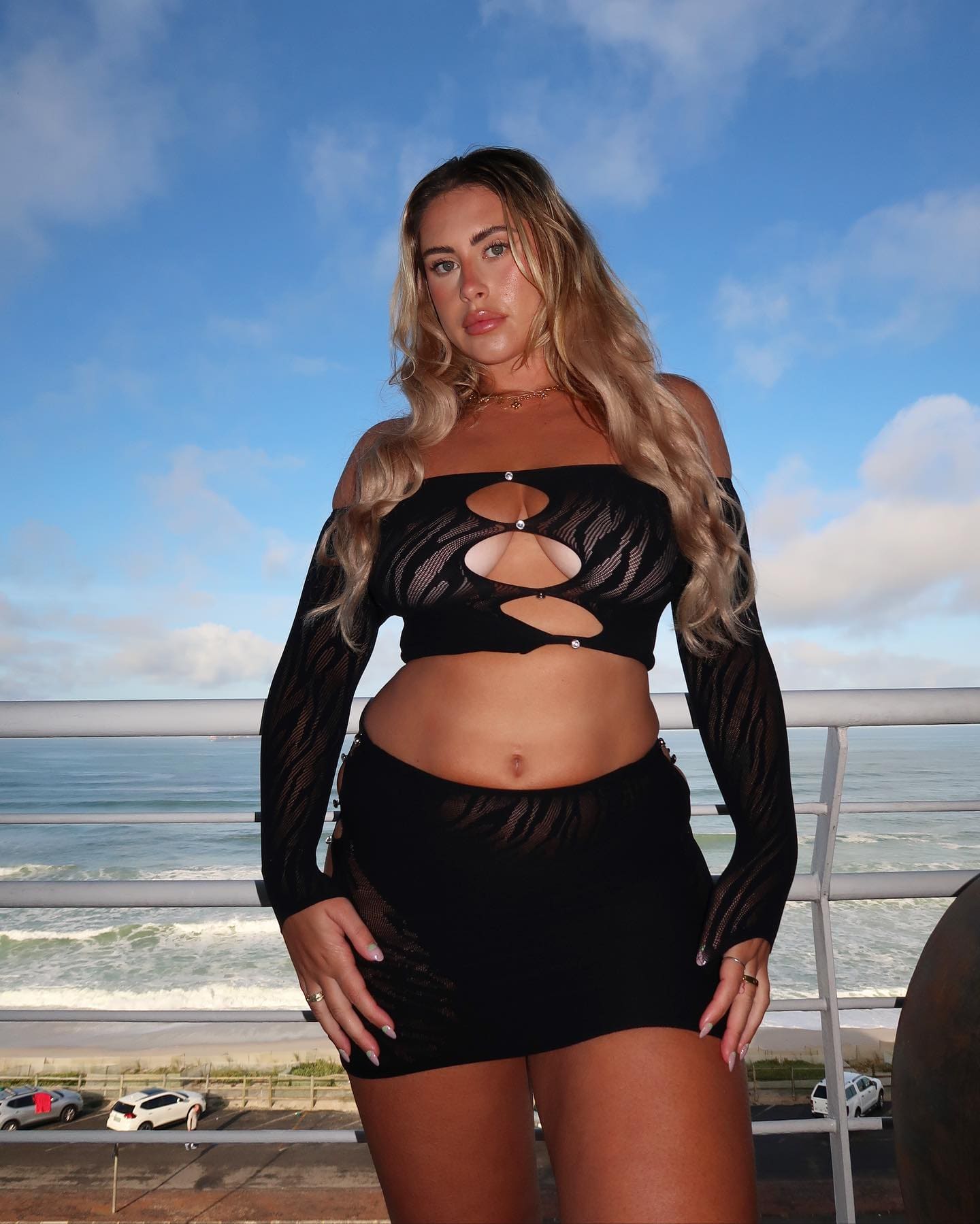 Blonde woman in black cut-out mesh top and skirt stands on a balcony with ocean view, hands on hips.