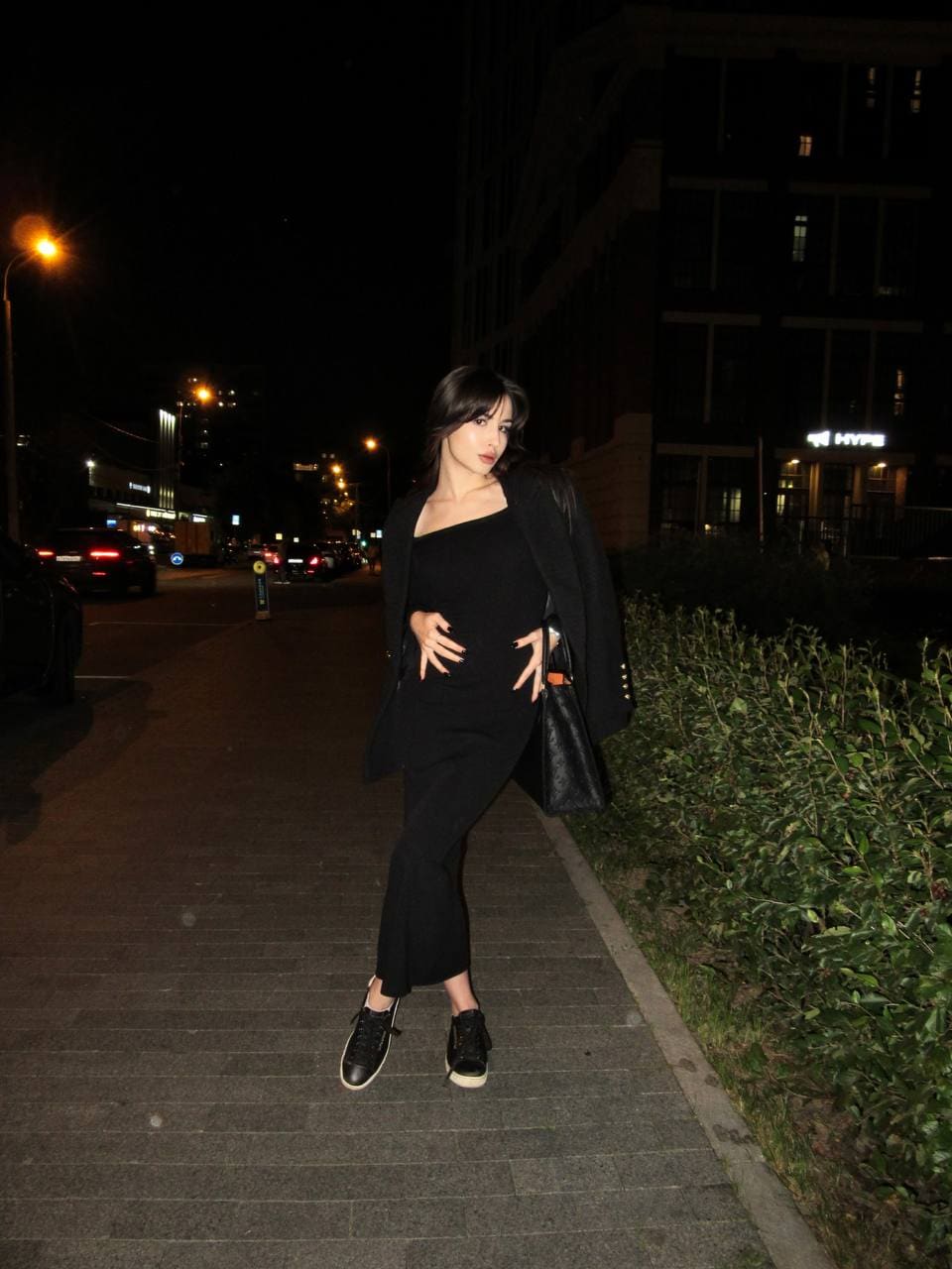 A woman stands on a night sidewalk in a black one-shoulder dress, blazer, and sneakers, holding a handbag.