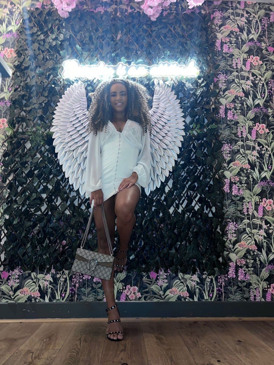 A woman stands before a floral wall wearing white angel wings, a white short dress, black strappy heels, and holding a patterned handbag.
