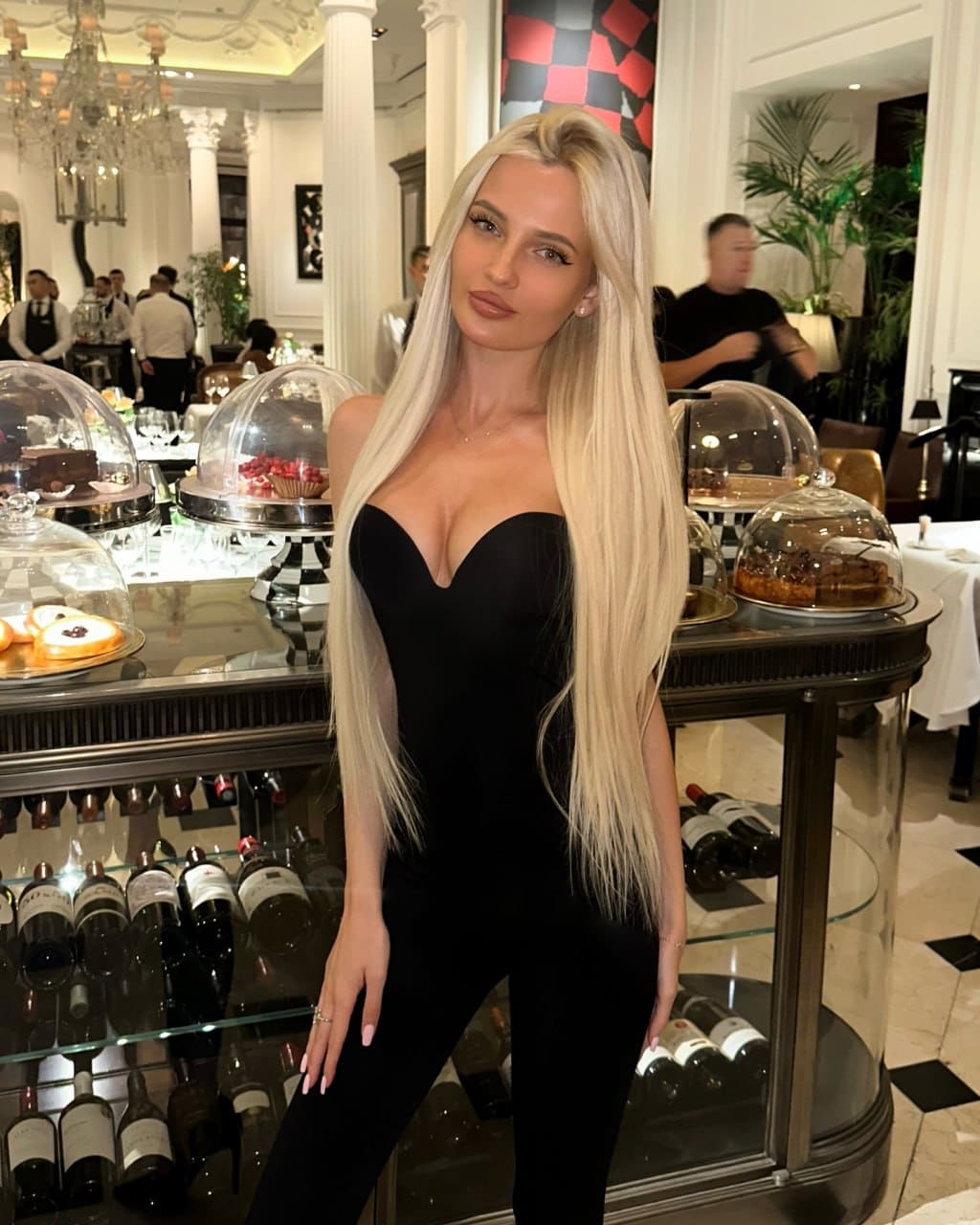 A woman stands in a formal dining room wearing a black dress with a sweetheart neckline, posing in front of a glass display case.