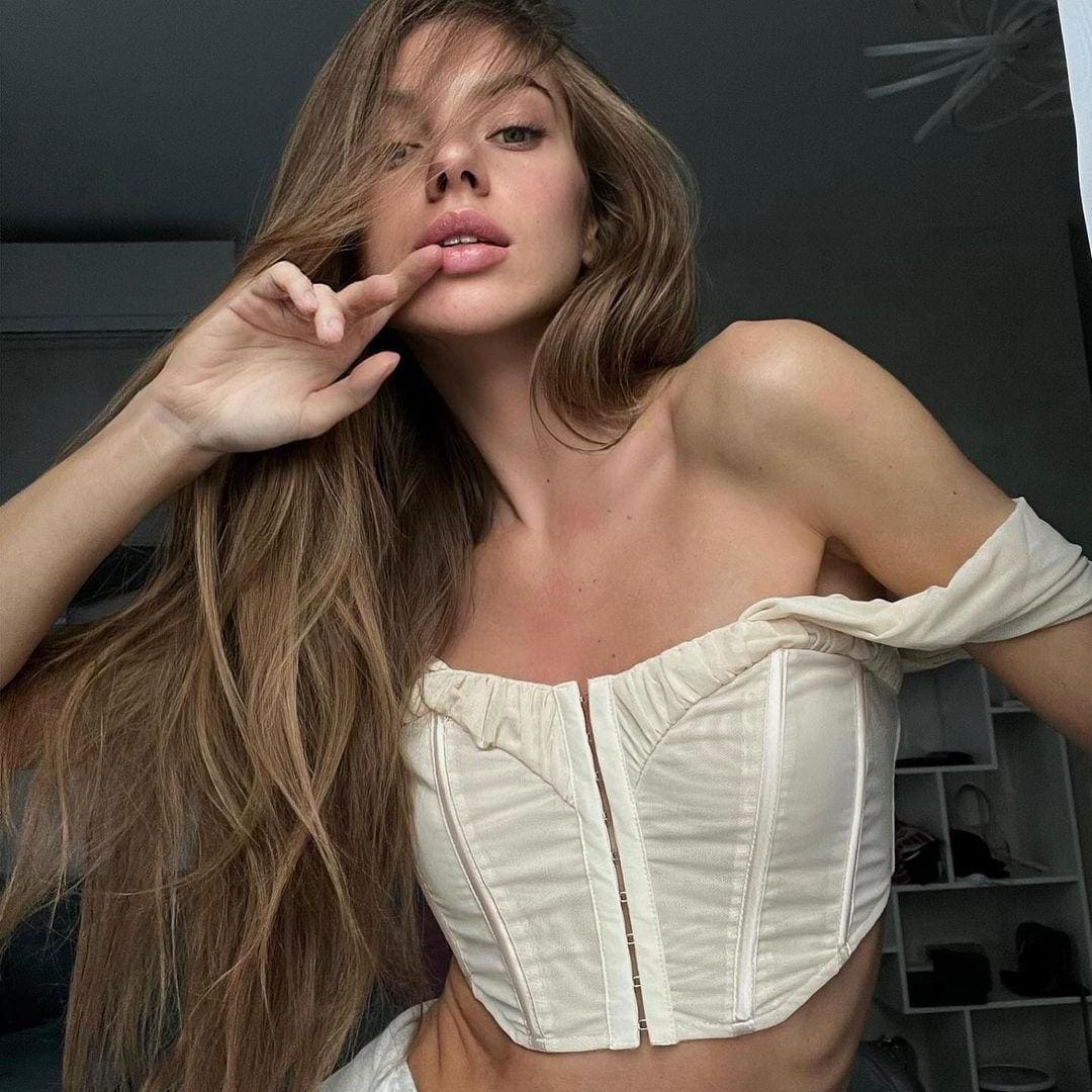 A woman with wavy hair wears a white strapless corset top with an off-shoulder sleeve, hand near mouth, against a dark wall with white shelving.