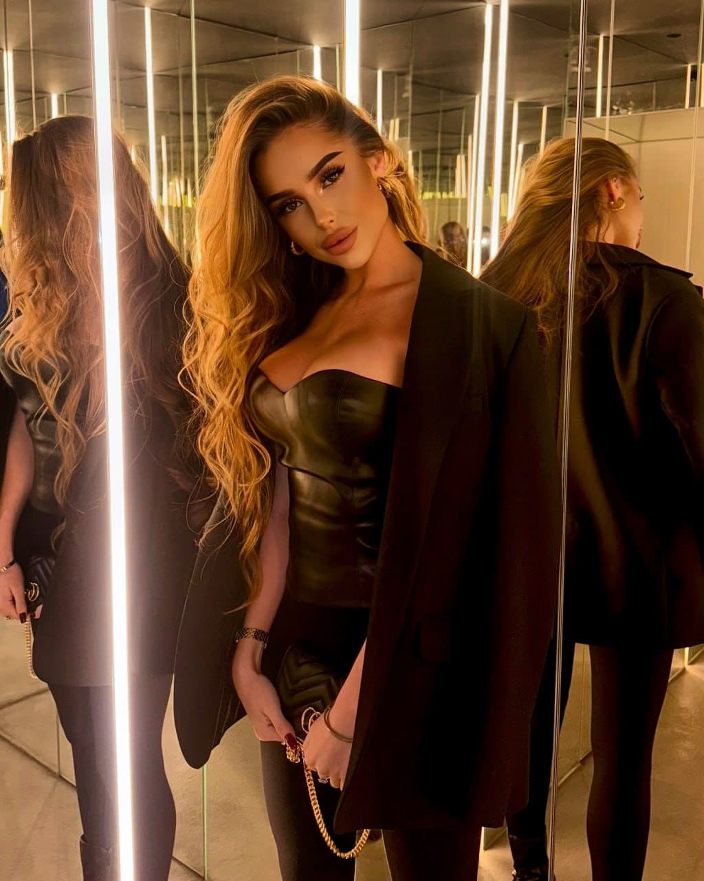 In a mirrored room with light strips, she wears a black leather bustier under an open blazer.