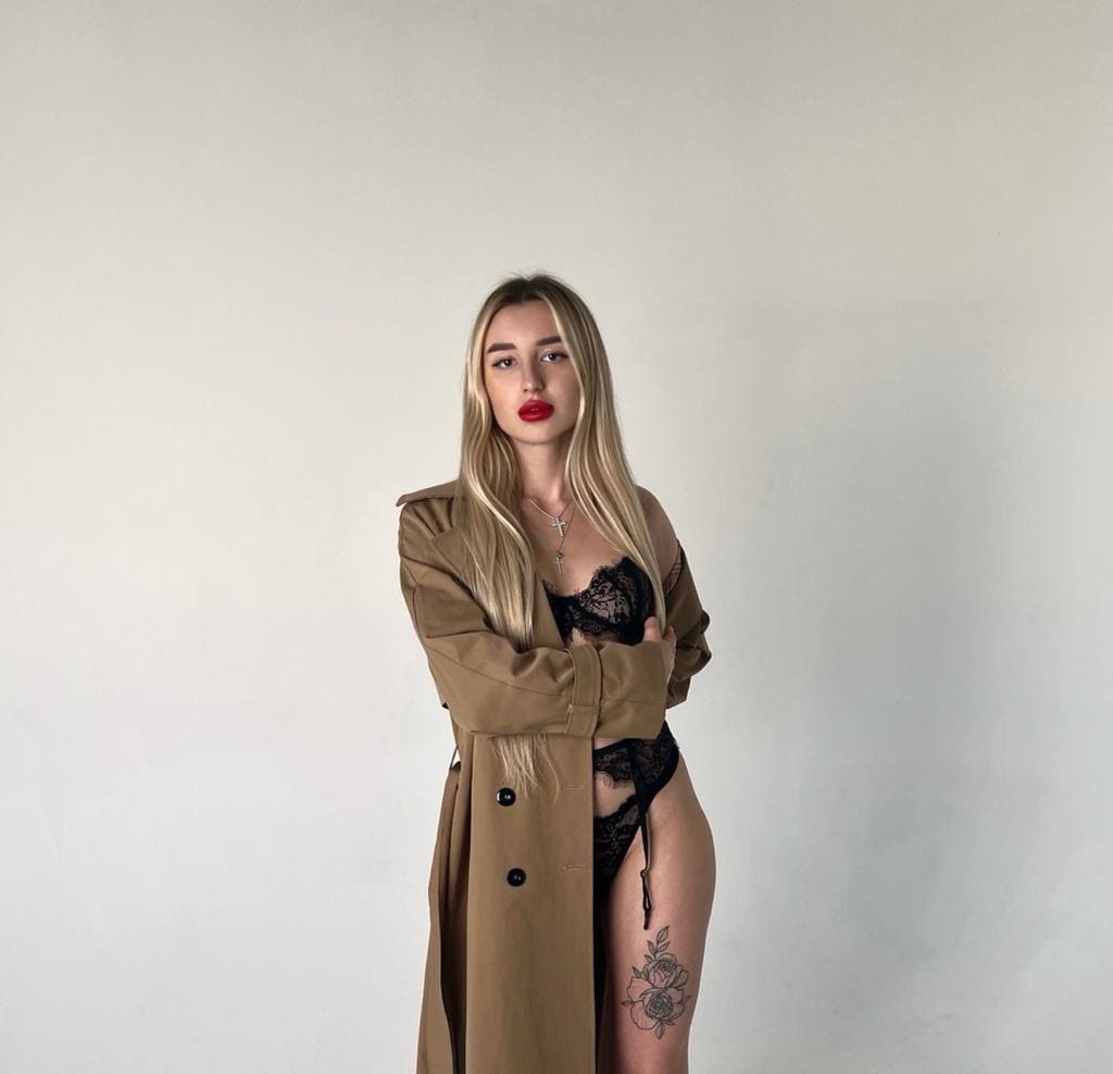 Charlene stands with arms crossed in an open tan trench coat and black lace bodysuit, showing a thigh tattoo against a white wall.