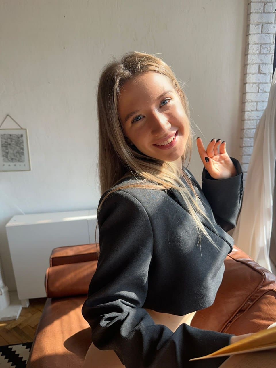 Yvonne sits on a brown leather sofa in a sunlit room, wearing a dark blazer and raising a hand with black nails toward the camera.