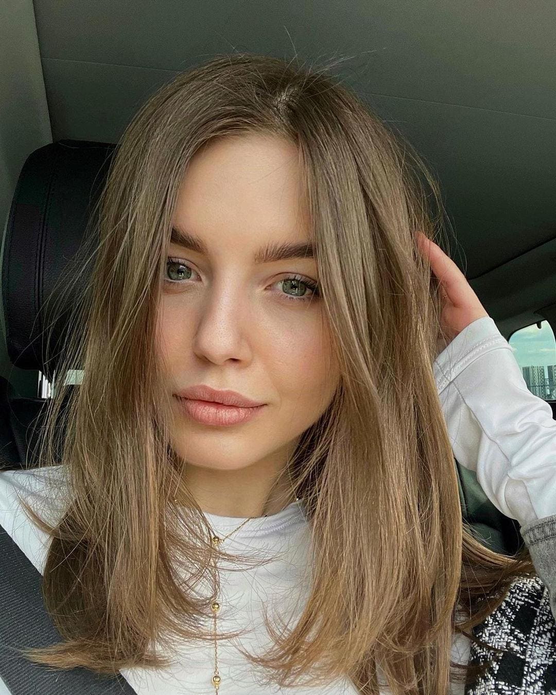Faith sits in a car wearing a white long-sleeved top and gold necklace, touching her long light-brown hair.