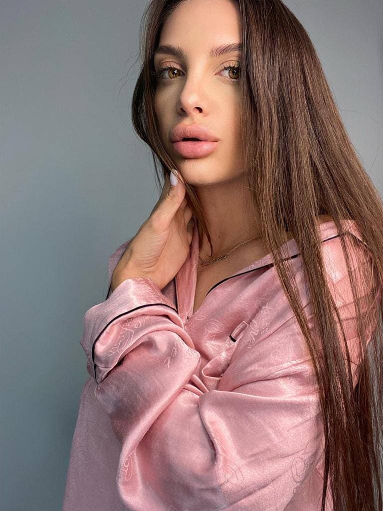 Leila poses in a pink satin pajama top with black piping, hand near her chin, against a plain gray background.