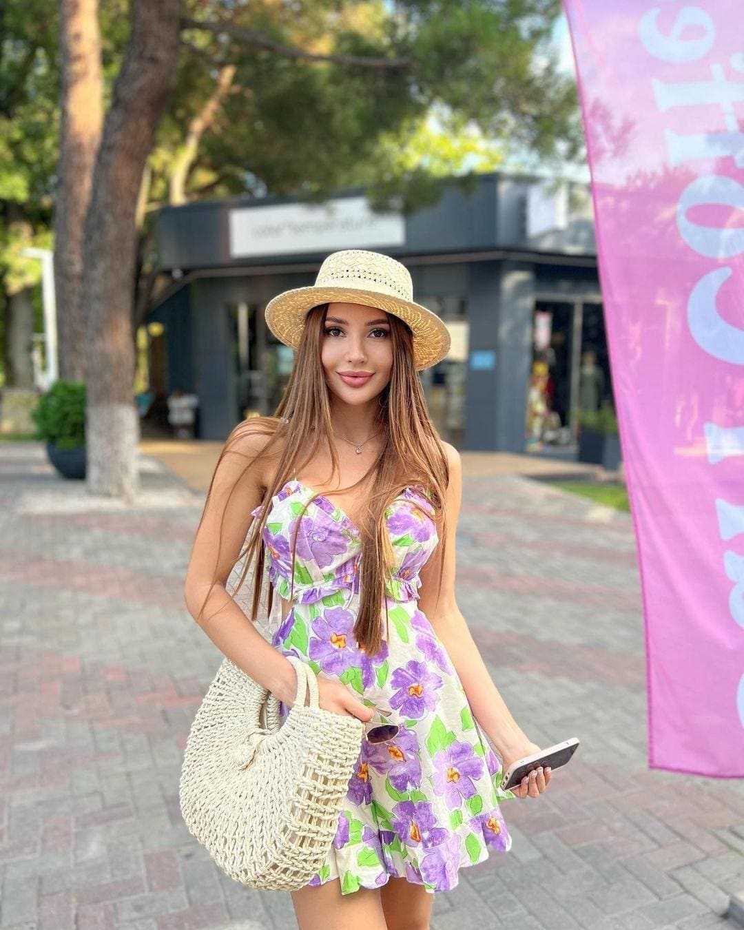 A woman in a straw hat and floral dress stands on an outdoor walkway, holding a woven bag and phone near trees.