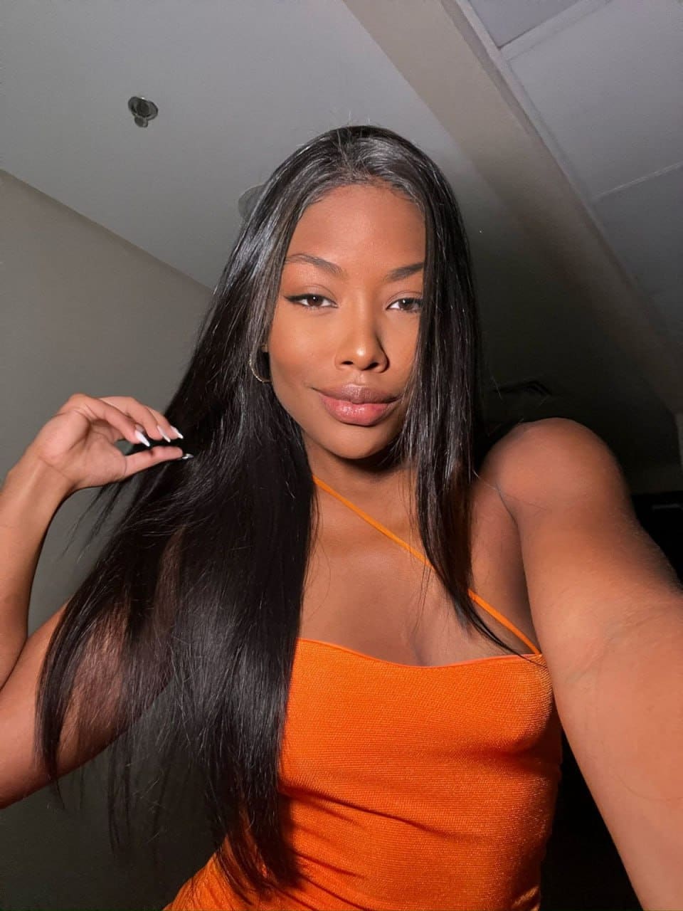 Selfie of a woman in an orange spaghetti-strap top holding a strand of her long black hair indoors.