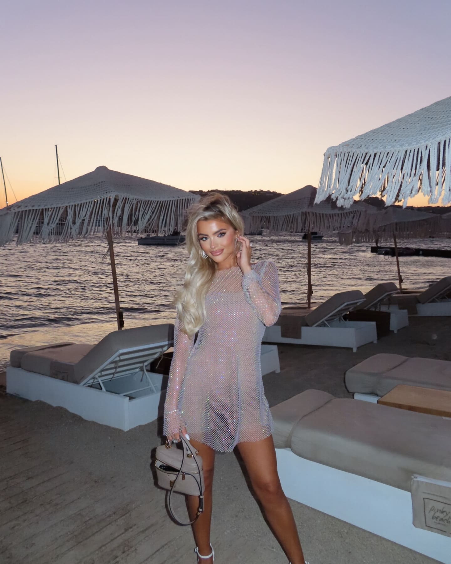 A woman in a sheer sparkly mini dress holds a handbag on a sandy beach at dusk, with umbrellas and chairs behind.