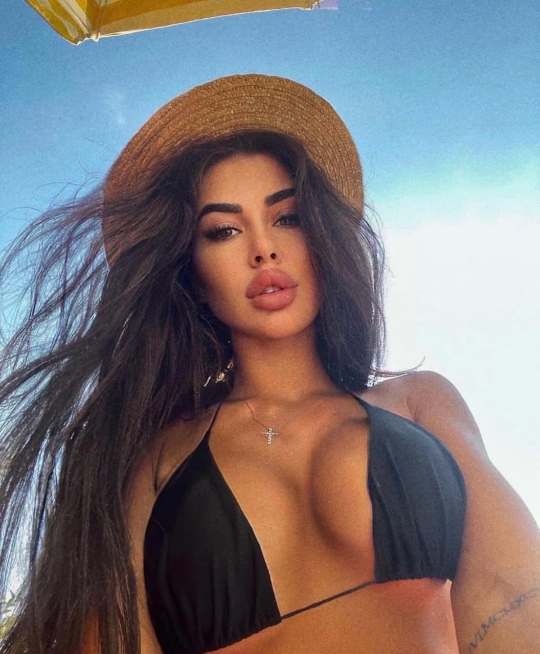 Woman wearing a straw hat and black halter bikini top poses outdoors under a blue sky, with the edge of a yellow umbrella visible above her.