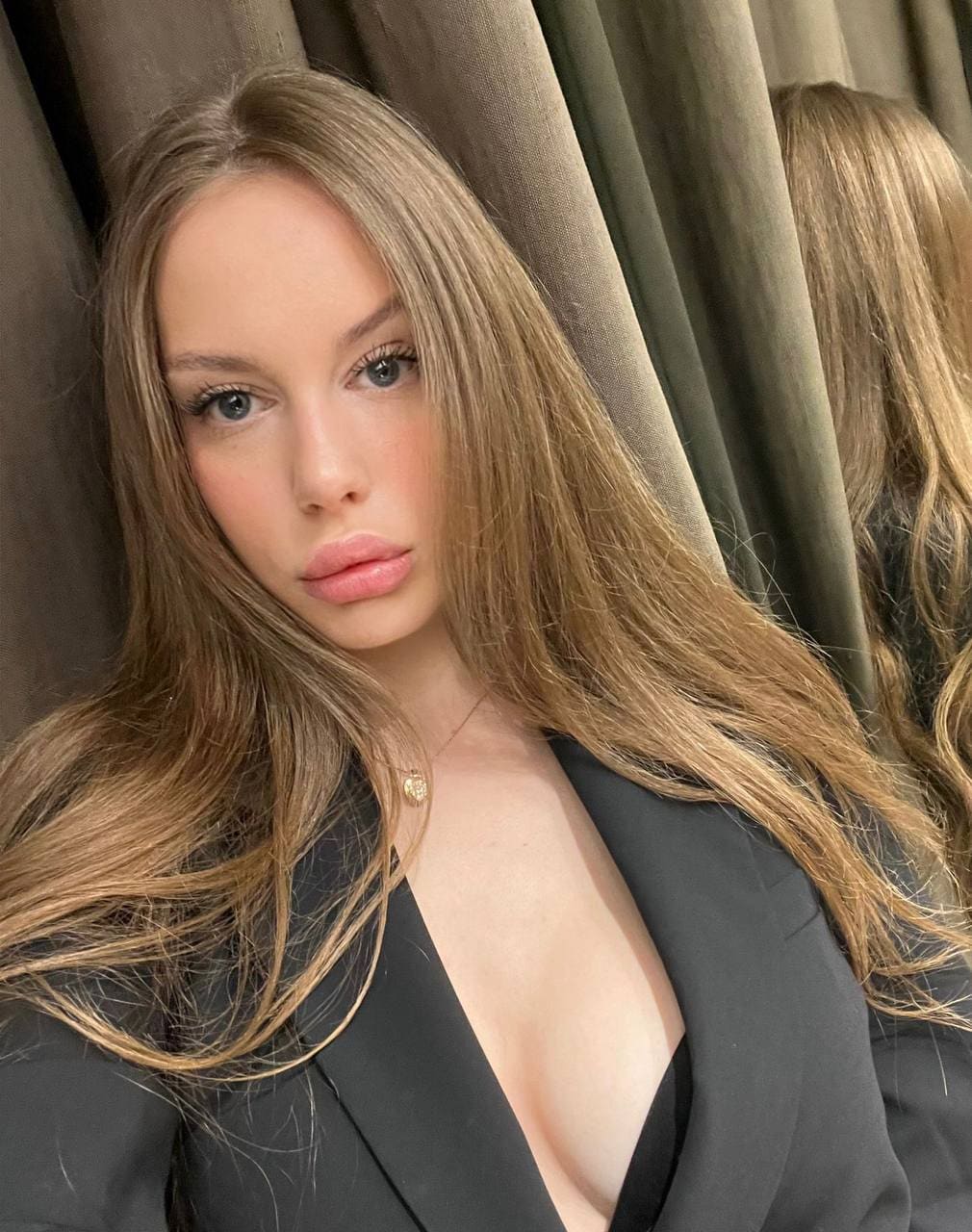 A woman in a black blazer with a deep V-neckline poses before a mirror, with olive-green curtains in the background.