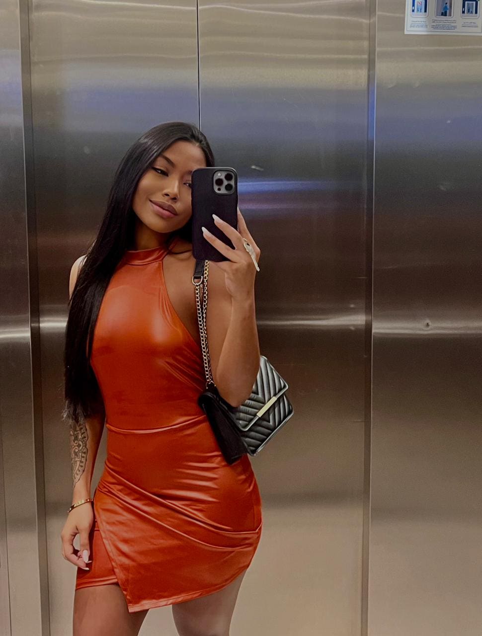 Mirror selfie in an elevator showing a woman in an orange mini dress holding a phone and black shoulder bag.
