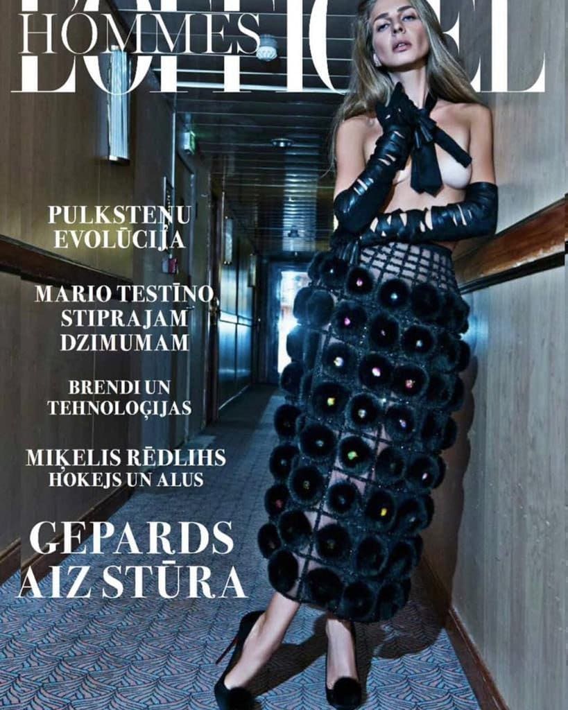 A woman in a black halter top, leather gloves, and pom-pom skirt stands in a hotel corridor wearing heels.