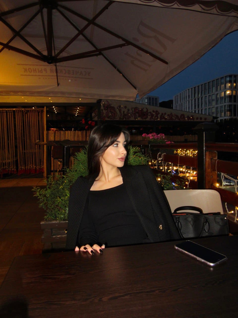A woman sits at a wooden table under a patio umbrella, wearing a black top and blazer, with a phone and bag.