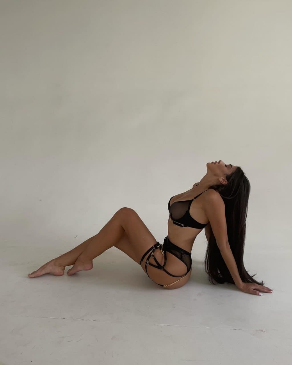 Woman sits on a light floor against a plain wall in a black sheer bra and strappy underwear with gold chains, head tilted back and one arm extended behind.