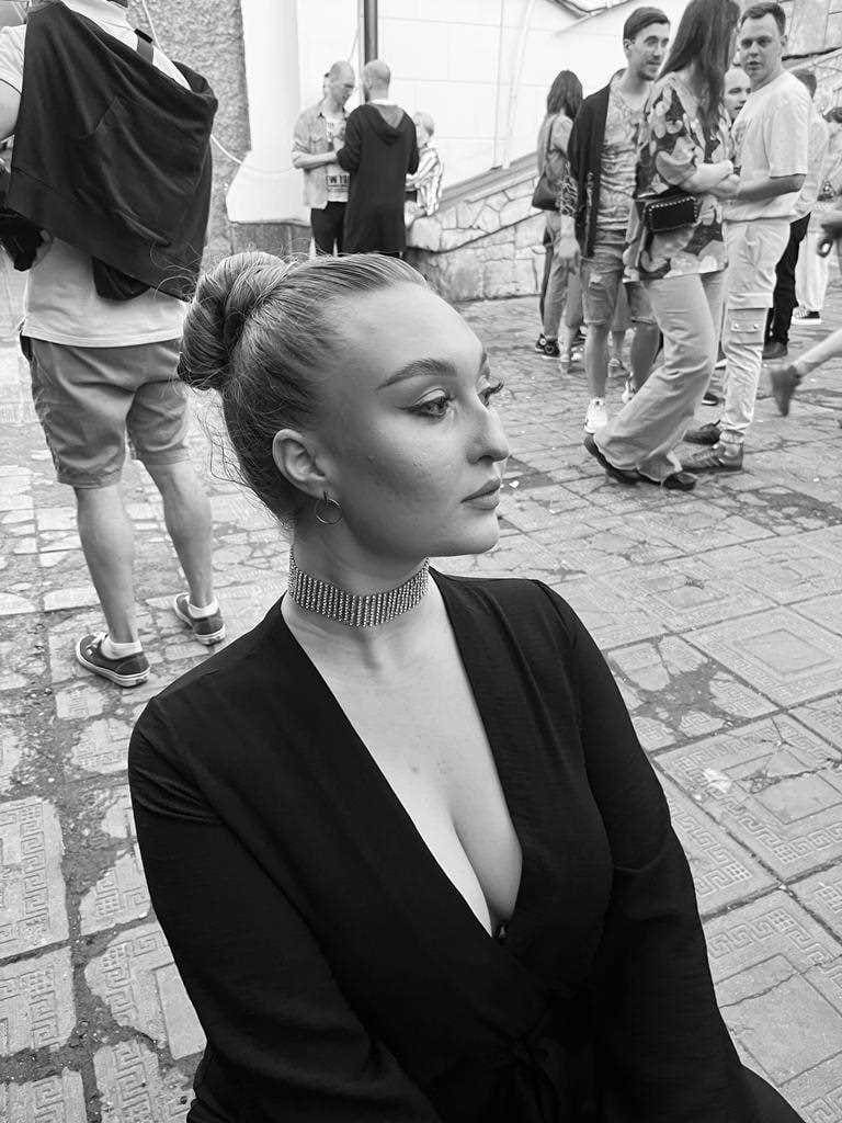 Woman with a bun sits outdoors on pavement wearing a black V-neck top and beaded choker, with people standing in the background.