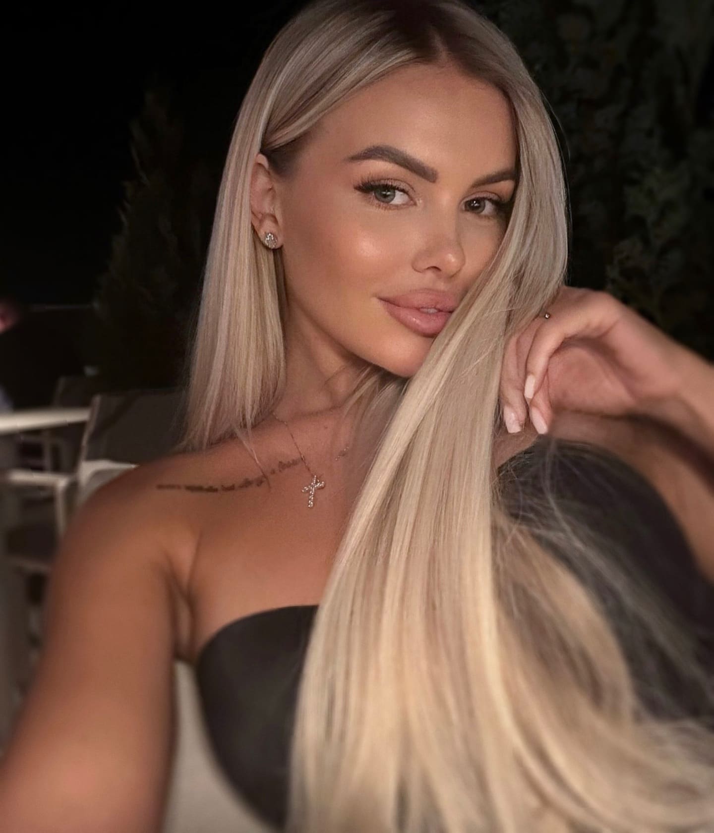Mirshi with long straight blonde hair wears a dark strapless top and cross necklace, holding a hair strand near her face in a dimly lit outdoor setting.