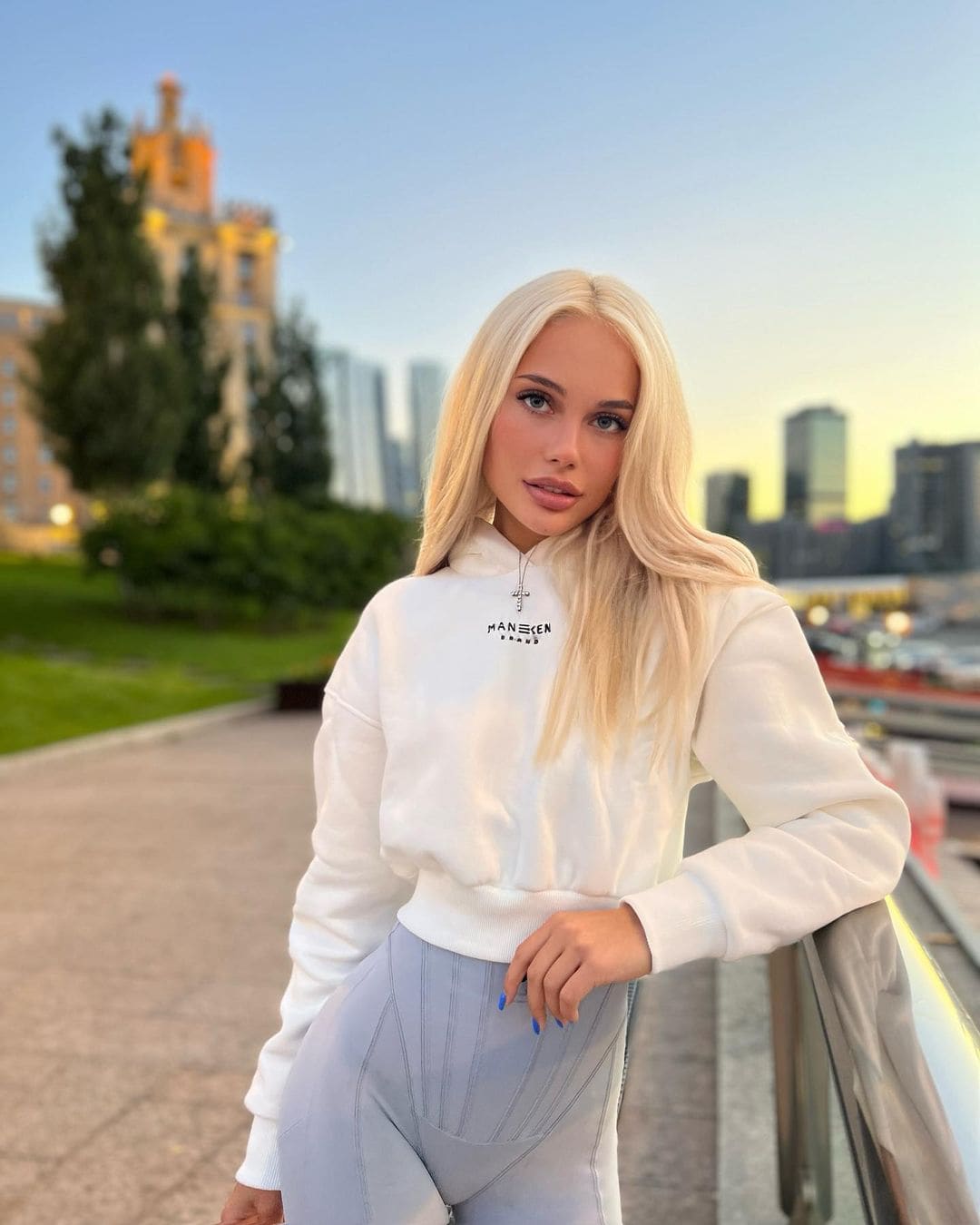 A blonde woman in a white hoodie and grey pants stands on a walkway at dusk, city skyline behind her.