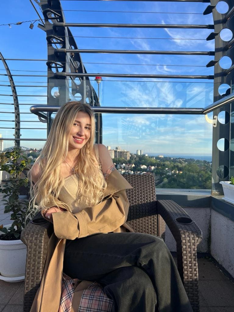 A blonde woman sits in a woven armchair on a rooftop terrace wearing a beige tank top and tan jacket, with a cityscape behind her.