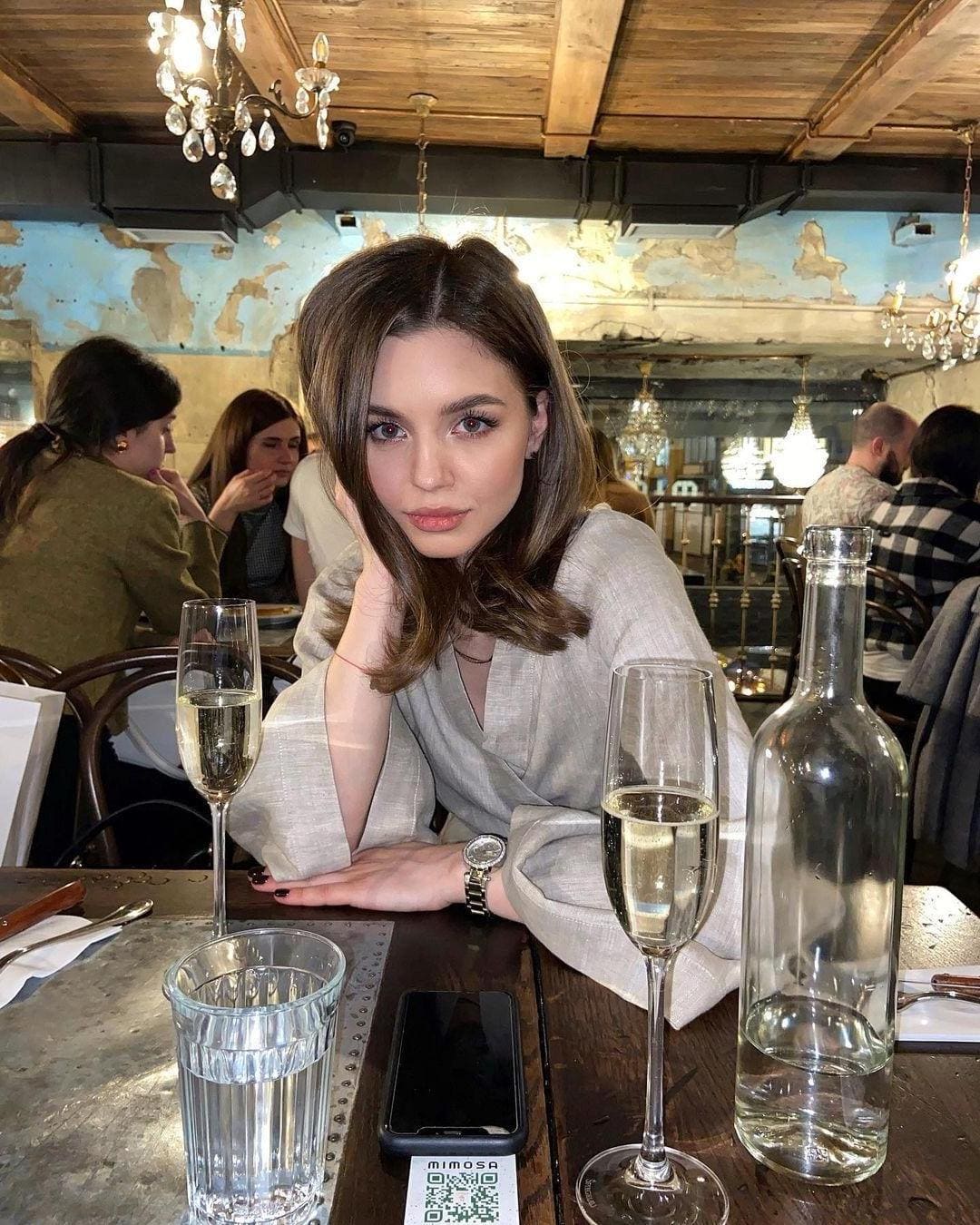 Woman in a grey wrap top sits at a restaurant table with champagne flutes and water, under chandeliers.