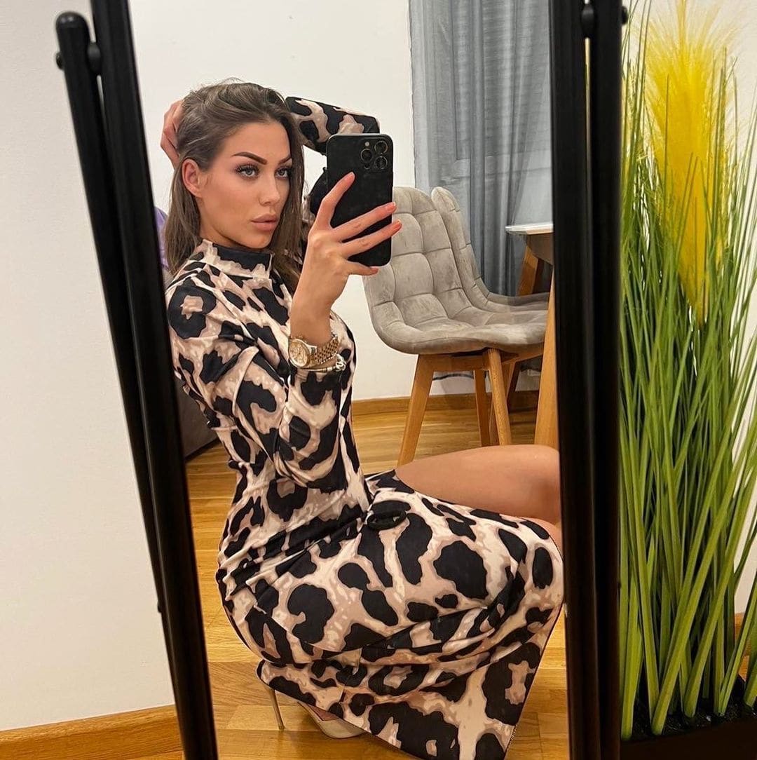 A woman in an animal print dress sits on a wooden floor, holding a phone near a mirror.
