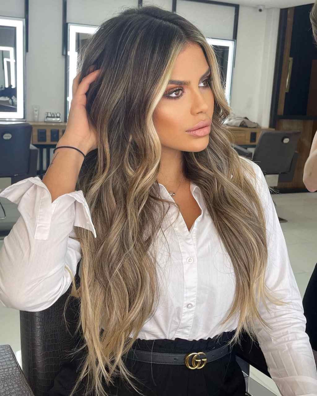 Seated in a salon chair, she wears a white button-up shirt with rolled sleeves and a black belt, touching her hair.