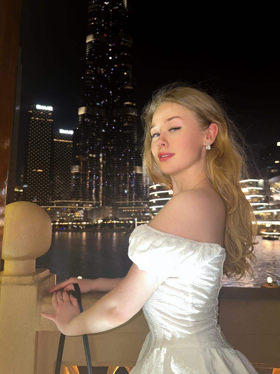 Lira stands at a stone railing at night in a white off-the-shoulder gown, with a lit city skyline and water behind her.