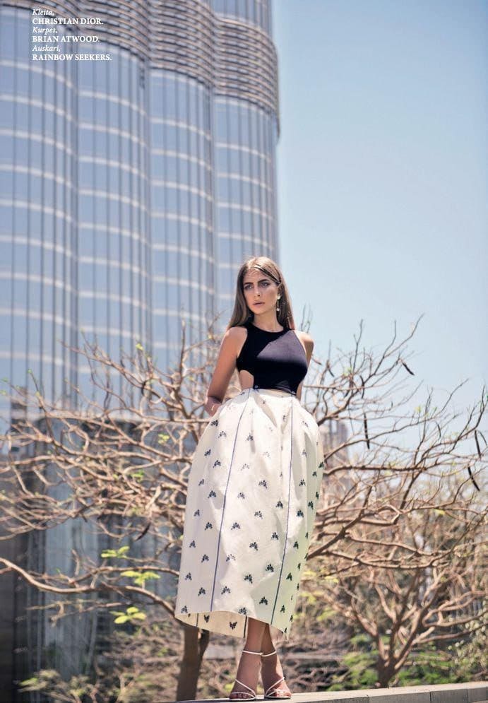 A woman in a black cutout top and white patterned skirt stands outdoors near a glass building and bare branches.