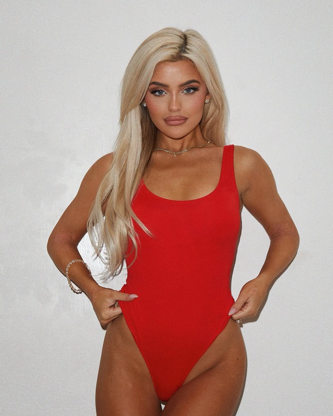 Avil stands with hands on hips against a light background, wearing a red sleeveless one-piece swimsuit.