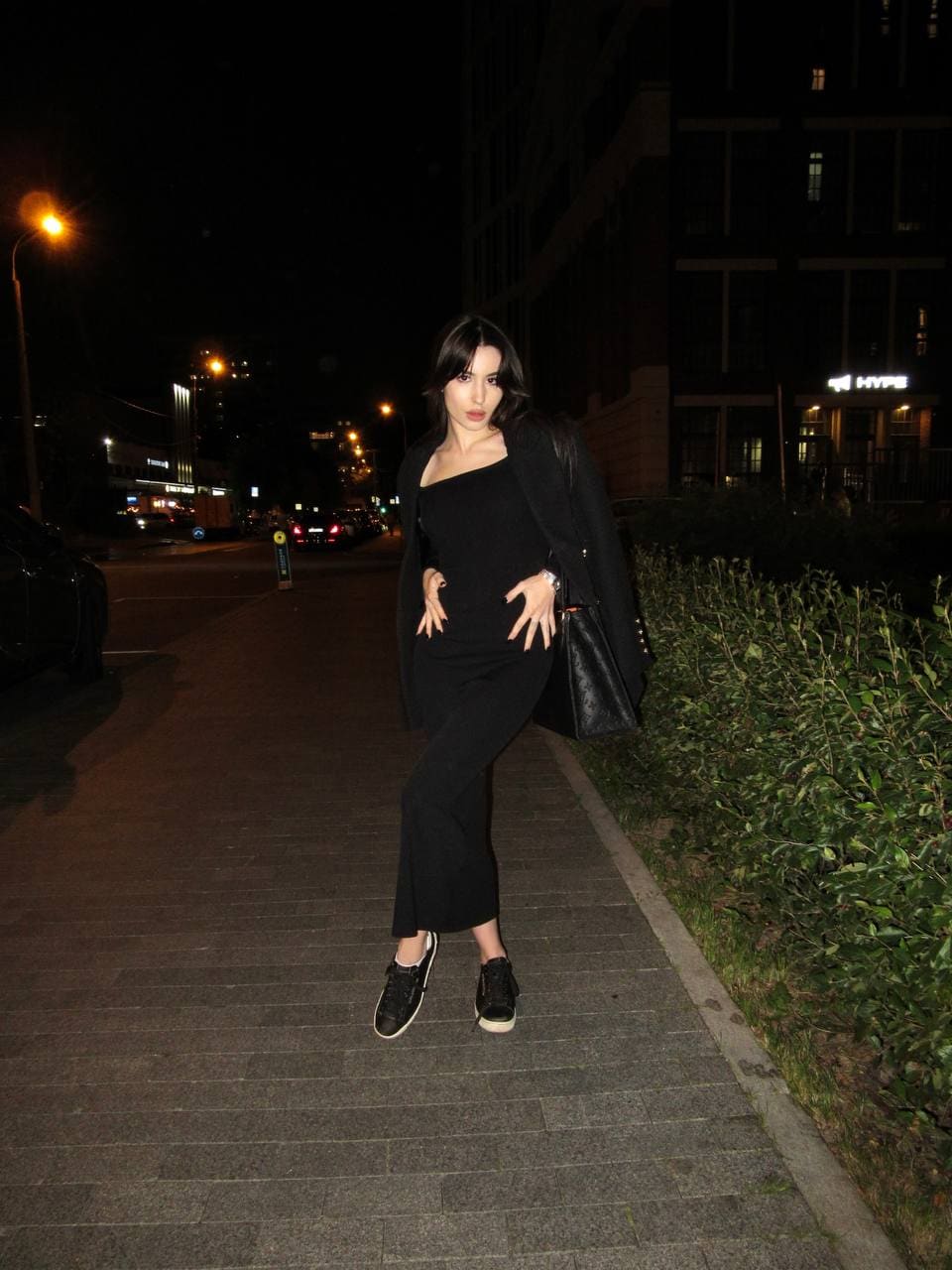 Night sidewalk scene: a woman in a black one-shoulder dress, open coat, and sneakers, hand on hip.