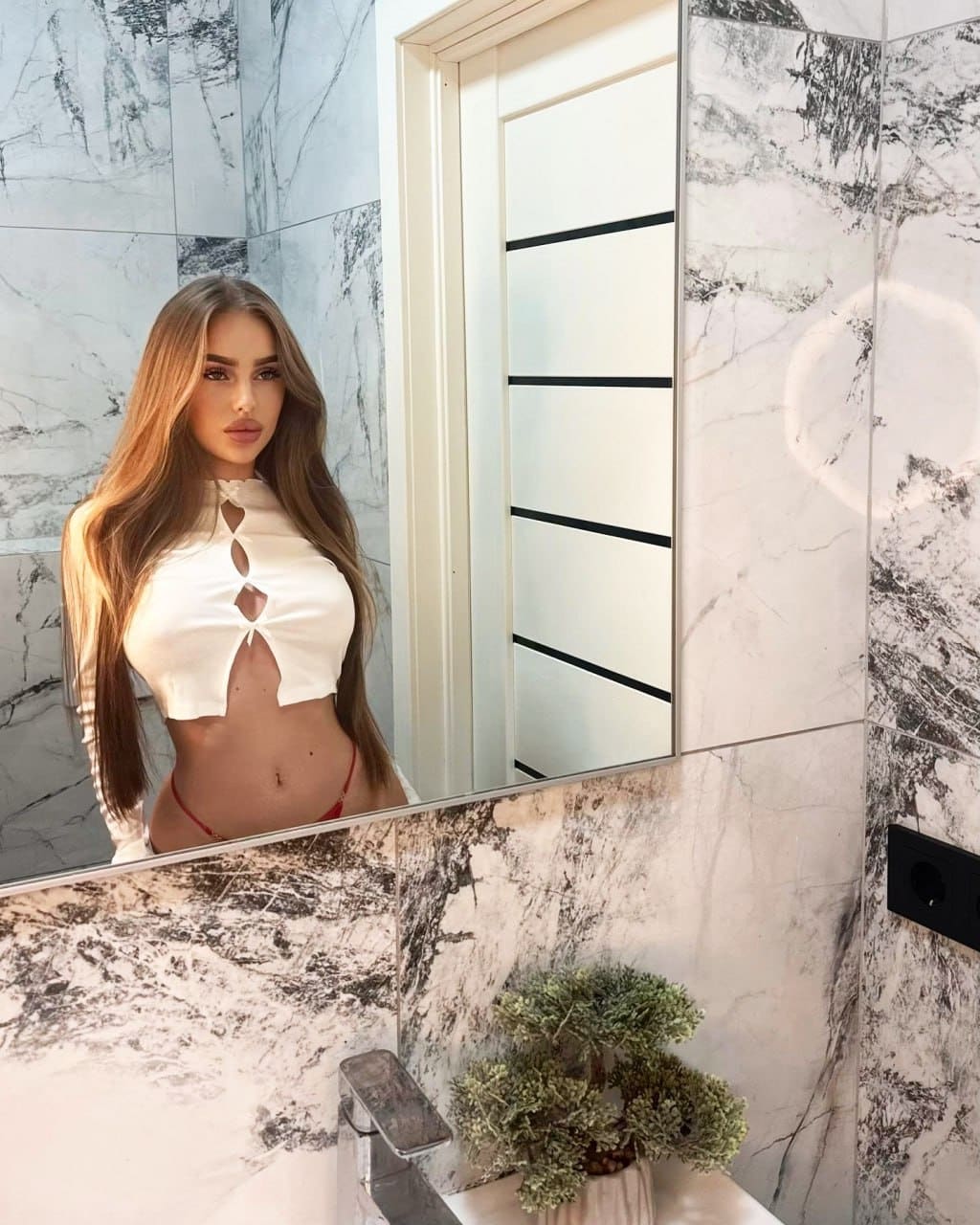 Facing a mirror in a marble bathroom, she wears a white keyhole crop top and red bikini bottoms.