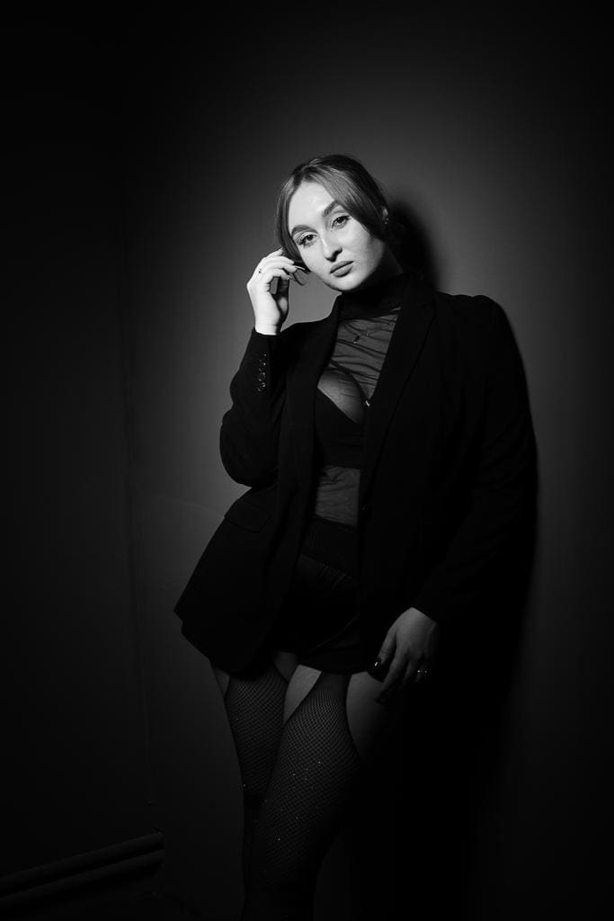 Woman stands in a dark studio wearing a black blazer, sheer mesh top, and fishnets, with one hand raised near her face.