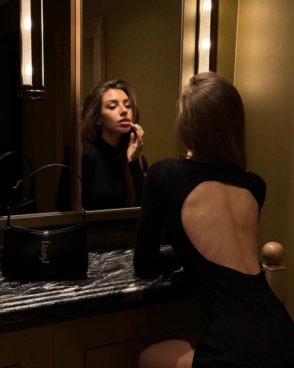 Seen from behind, a woman sits at a marble vanity in a black open-back top, her reflection touching lips, with a black handbag on the counter.