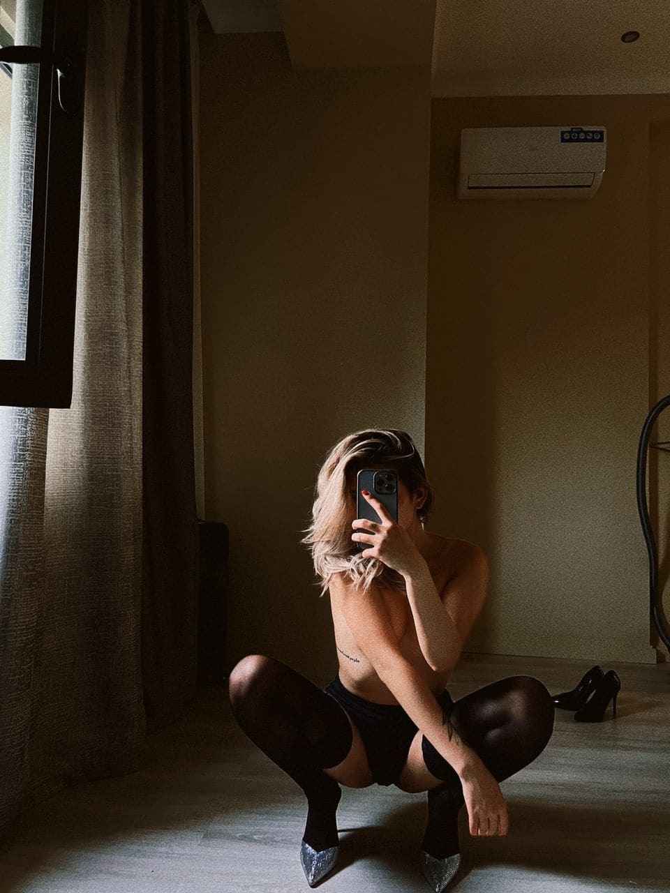 A blonde person crouches on a carpeted floor in a dim room, wearing black shorts, sheer stockings, and high heels.