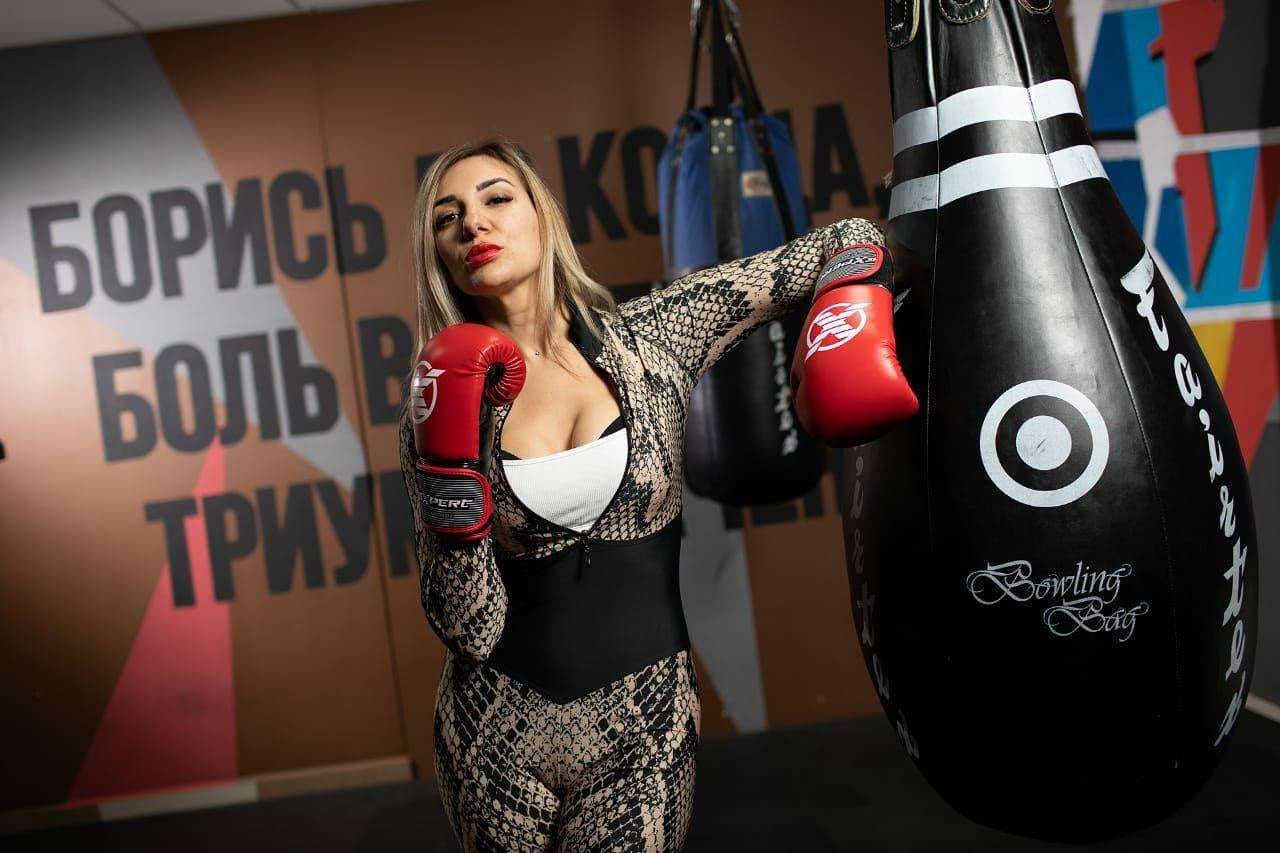 Alina stands in a gym wearing a snakeskin bodysuit and red boxing gloves, posing beside a black heavy bag with Cyrillic text on the wall.